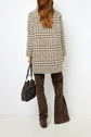A woman with long brown hair, wearing a manteau court à carreaux in a black-and-white houndstooth pattern and brown patterned pants, holds a black handbag. She is standing against a plain white wall, photographed from the back.
