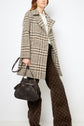 A woman wearing a manteau court à carreaux in beige and brown houndstooth, a cream blouse, brown patterned wide-leg pants, and carrying a dark brown handbag stands against a white background.