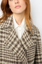 A woman with light red hair wears a manteau court à carreaux in brown and cream plaid over a white collared shirt, facing the camera with a neutral expression.