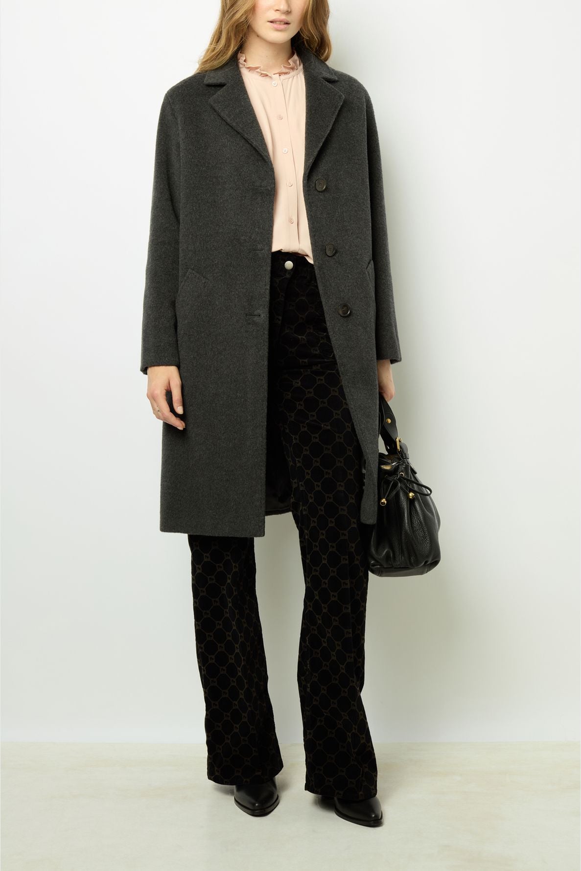 Woman wearing a dark gray manteau mi-long over a pale pink blouse and black patterned pants, holding a black handbag, standing against a plain light background.