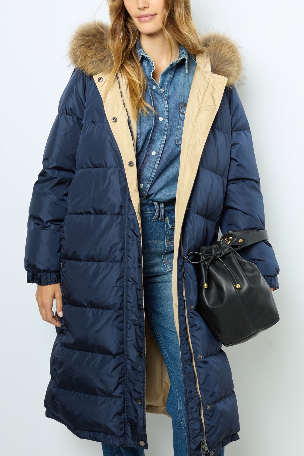 Woman wearing a long navy doudoune à capuche with a fur-trimmed hood over a denim outfit, holding a black drawstring bag.