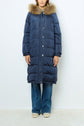 A person wearing a long, navy blue doudoune à capuche with a furry hood, blue jeans, and beige shoes stands against a plain white background.