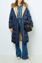 A woman wears a long navy puffer coat, or doudoune longue, with a fur-trimmed hood—perfectly blending style and warmth. She pairs her doudoune à capuche with blue jeans, a denim shirt, and white sneakers, carrying a small black drawstring bag.
