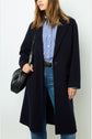 Woman in a navy manteau droit over a blue-and-white striped shirt and jeans, carrying a black shoulder bag. Her hair is loose and she is standing against a plain light background.