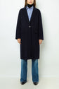 A person wearing a dark navy manteau droit knee-length coat over a blue and white striped shirt, blue jeans, and black shoes, standing against a plain white background.