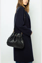A woman in a dark, long manteau droit carries a black leather shoulder bag with gold accents. The bag has a gathered top and an adjustable strap.