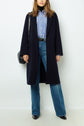Woman wearing a navy manteau droit over a blue striped shirt and blue jeans, black shoes, and carrying a black shoulder bag, standing against a plain white background.
