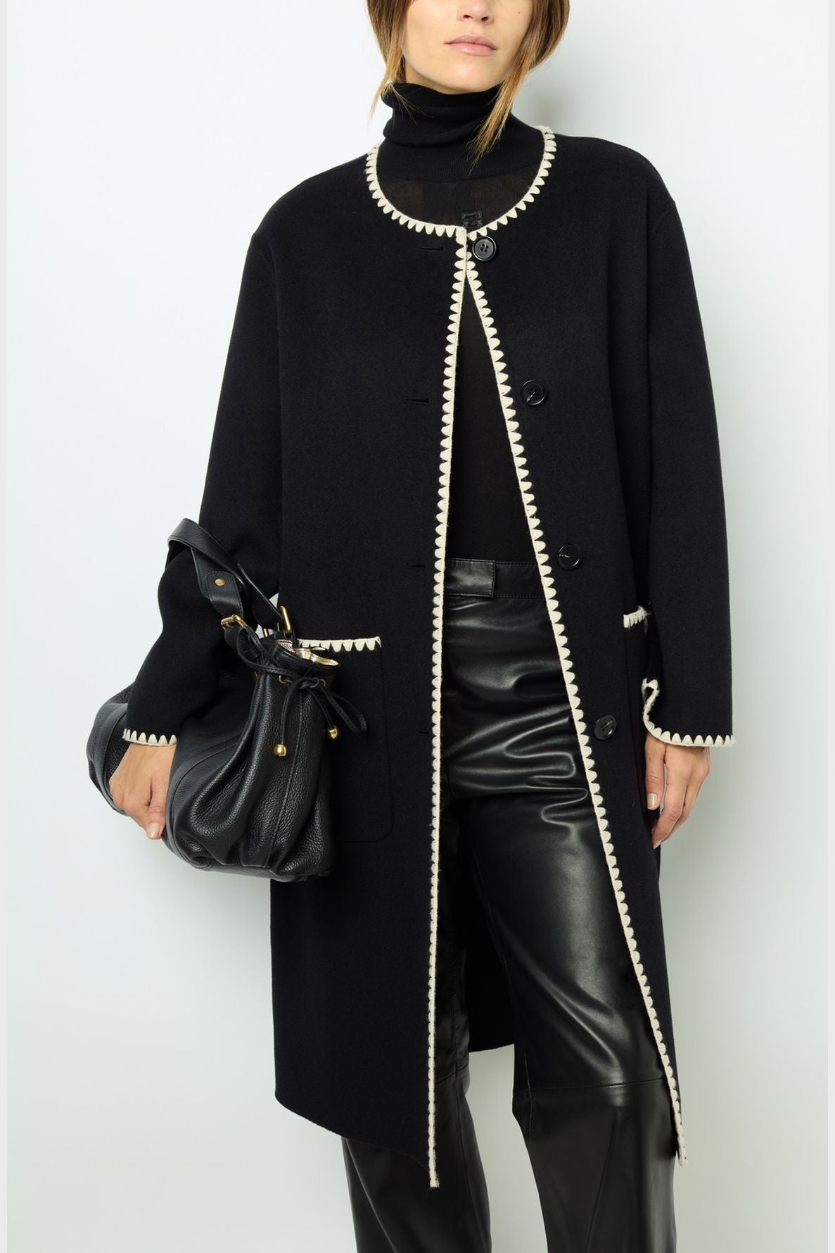 Woman wearing a manteau mi-long in black with white trim, a black turtleneck, black leather pants, and holding a black handbag, standing against a plain white background for a look sophistiqué.