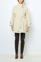A person wearing a cream-colored manteau mi-long ceinturé, crafted from soft laine alpaga, dark pants, and brown ankle boots stands against a plain white background. The coat features elegant button details at the neck and sleeves.