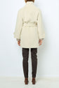 Person standing facing away from the camera, wearing a light beige manteau mi-long ceinturé in laine alpaga, dark pants, and brown heeled shoes, against a plain white background.