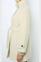 Woman in a beige manteau ceinturé with a high collar and buttoned cuffs, standing sideways against a plain light background. Crafted from laine alpaga, her manteau mi-long drapes elegantly as her face remains partially visible and one arm hangs by her side.