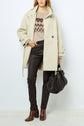 A person wearing a beige manteau ceinturé, a patterned sweater, dark pants, and brown boots holds a dark handbag. The outfit looks warm and stylish—perfect for fall or winter—especially with the chic manteau mi-long. The background is plain and light-colored.