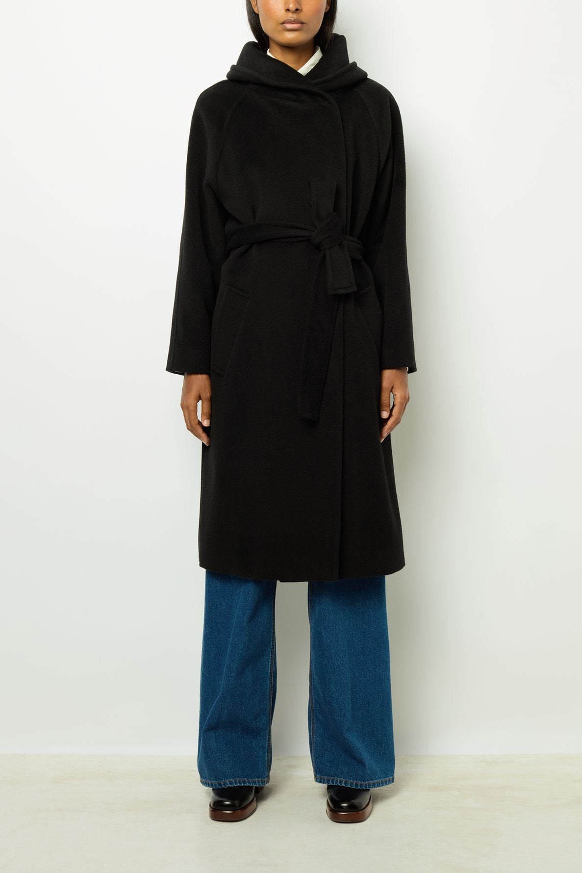 Person wearing a manteau long femme, a black belted coat reminiscent of a manteau laine alpaga, over blue wide-leg jeans and brown shoes, standing against a plain white background. Their head is partially cropped out of the image.