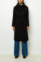 Person wearing a manteau long femme, a black belted coat reminiscent of a manteau laine alpaga, over blue wide-leg jeans and brown shoes, standing against a plain white background. Their head is partially cropped out of the image.