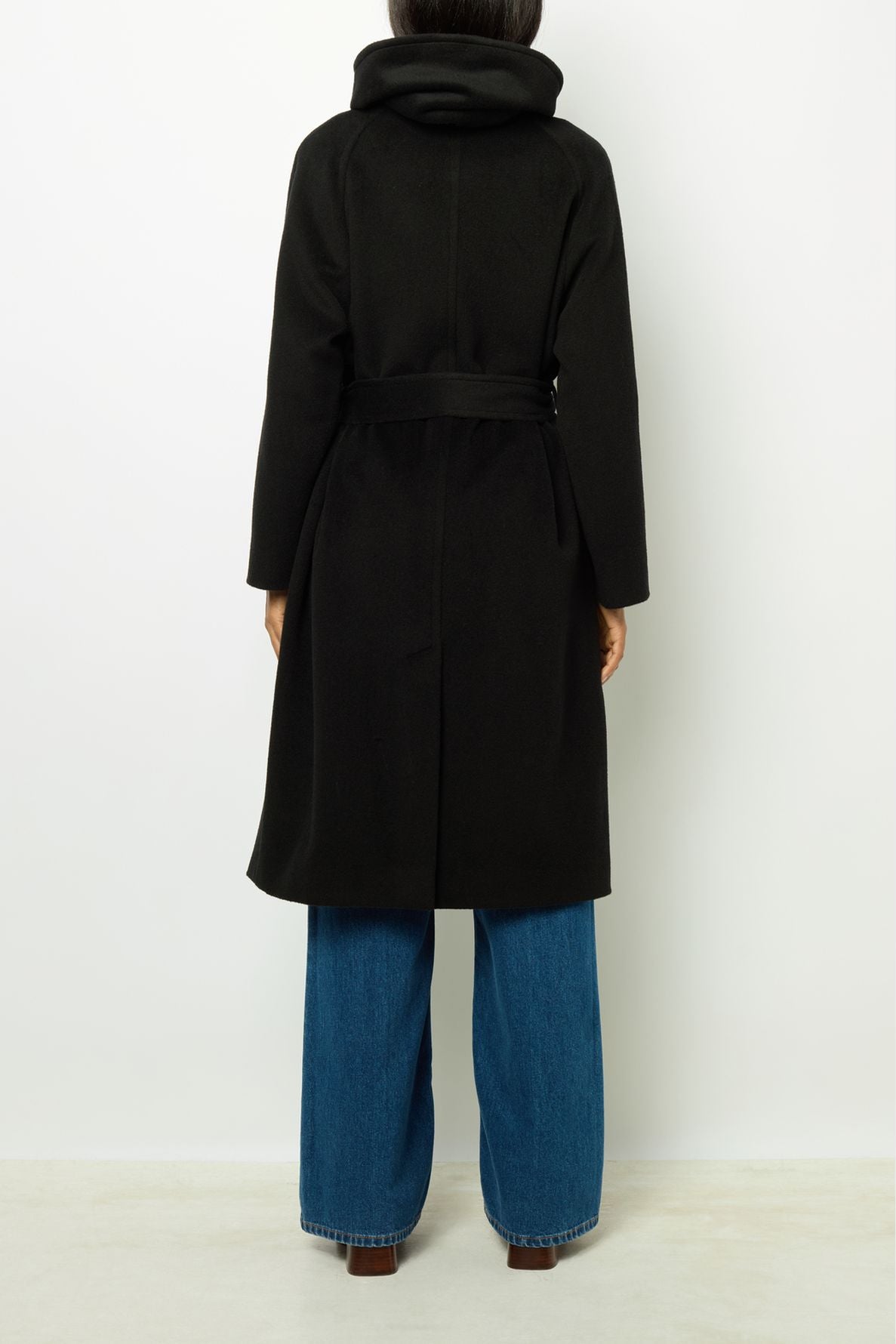 Person standing with their back to the camera, wearing a manteau long femme ceinturé in black, paired with wide blue jeans. The coat features a high collar and is crafted from luxurious laine alpaga, while the person’s hair is partially visible above the collar.