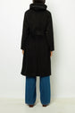 Person standing with their back to the camera, wearing a manteau long femme ceinturé in black, paired with wide blue jeans. The coat features a high collar and is crafted from luxurious laine alpaga, while the person’s hair is partially visible above the collar.