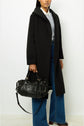 A person in a manteau long femme—an elegant black coat—paired with wide-leg blue jeans, holds a textured black handbag with gold hardware. The image is cropped at the upper chest, and the person stands against a plain white background.