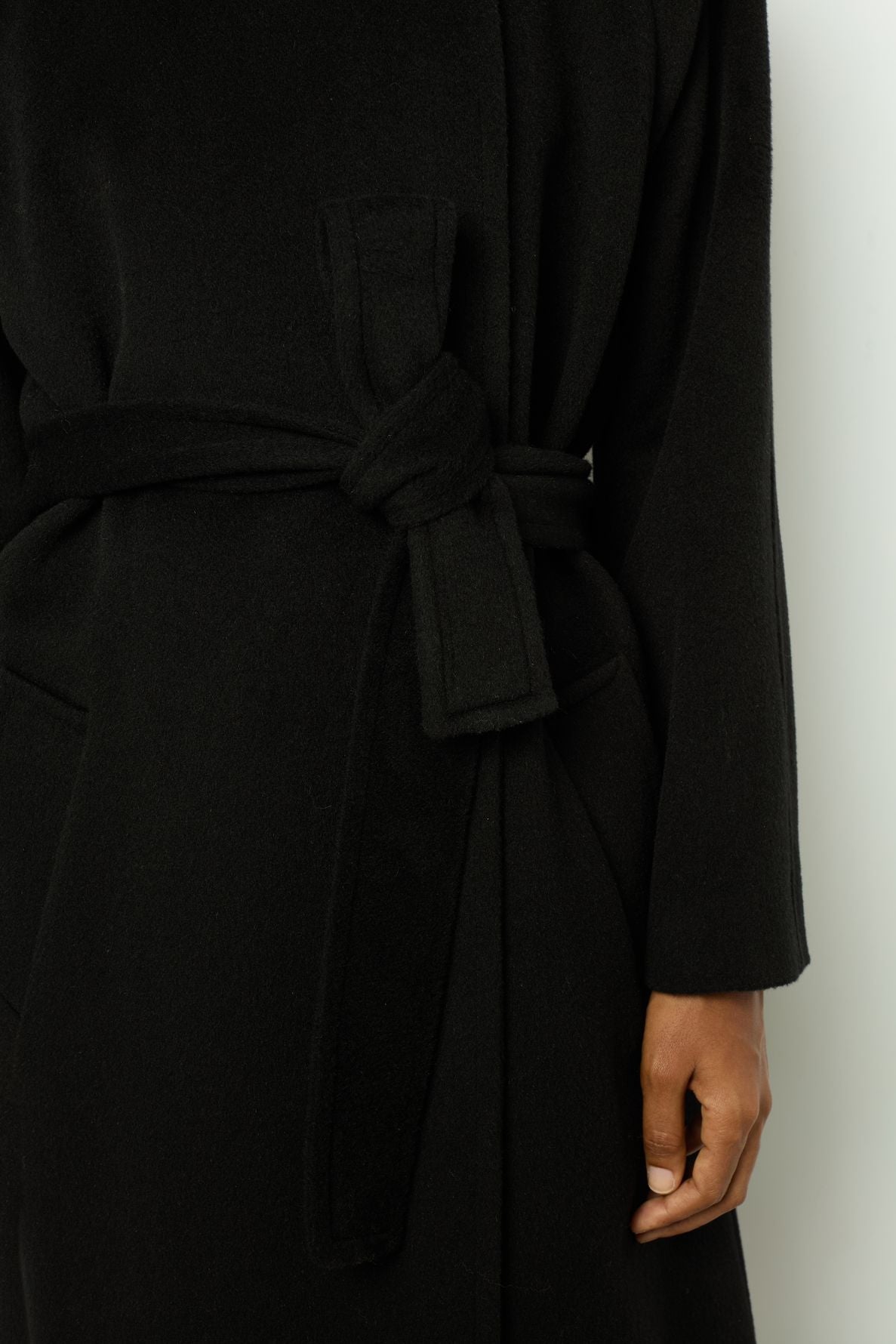 A person wearing a black manteau ceinturé, shown from the shoulders to mid-thigh. Their right hand is relaxed by their side, and the coat’s fabric belt is tied at the waist.