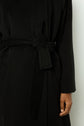 A person wearing a black manteau ceinturé, shown from the shoulders to mid-thigh. Their right hand is relaxed by their side, and the coat’s fabric belt is tied at the waist.