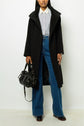 A person wearing a manteau long femme in black, layered over a light blue button-up shirt, high-waisted blue jeans, and brown shoes holds a black handbag. The image is cropped at the shoulders.
