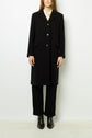 A person wearing a manteau mi-long femme in a long black coat over black pants and black boots stands against a plain white background. Their hands are at their sides, and their head is partially out of frame.