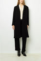 A woman stands against a white background wearing a manteau mi-long femme—a long black coat crafted from luxurious laine vierge—paired with a cream blouse, black pants, and black ankle boots. Her hair is loose and her hands rest by her sides.