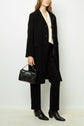 A woman wearing a manteau mi-long femme—a long black coat in the style of a manteau droit—over a cream blouse, black pants, and heeled boots, holds a black handbag with a chain strap while standing against a plain white background.