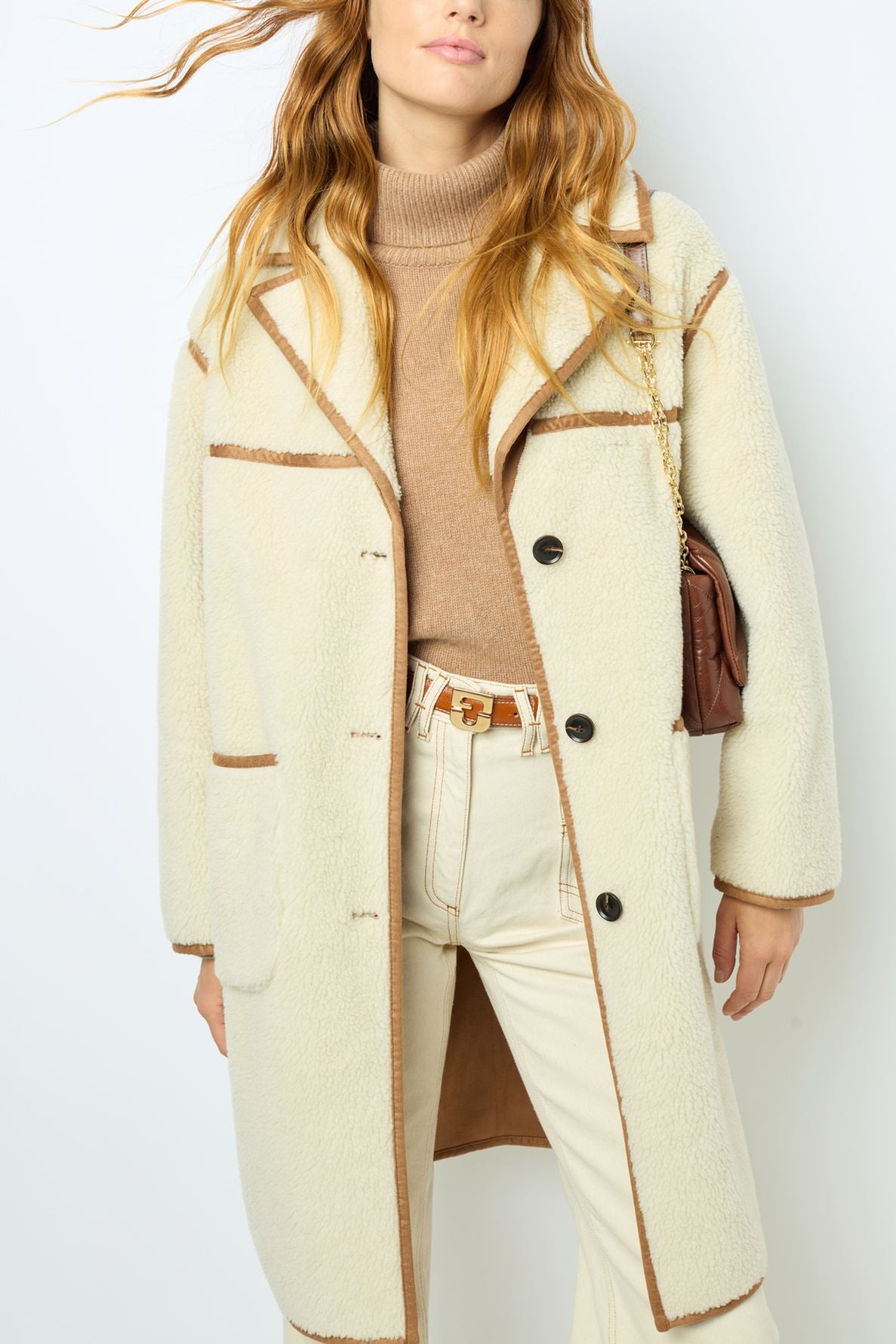 Woman wearing a cream manteau fausse fourrure with brown trim, a camel turtleneck sweater, cream pants, and a brown purse with a chain strap. Her hair is wavy and loose, and her face is partially out of frame—an effortlessly chic manteau hiver femme look.