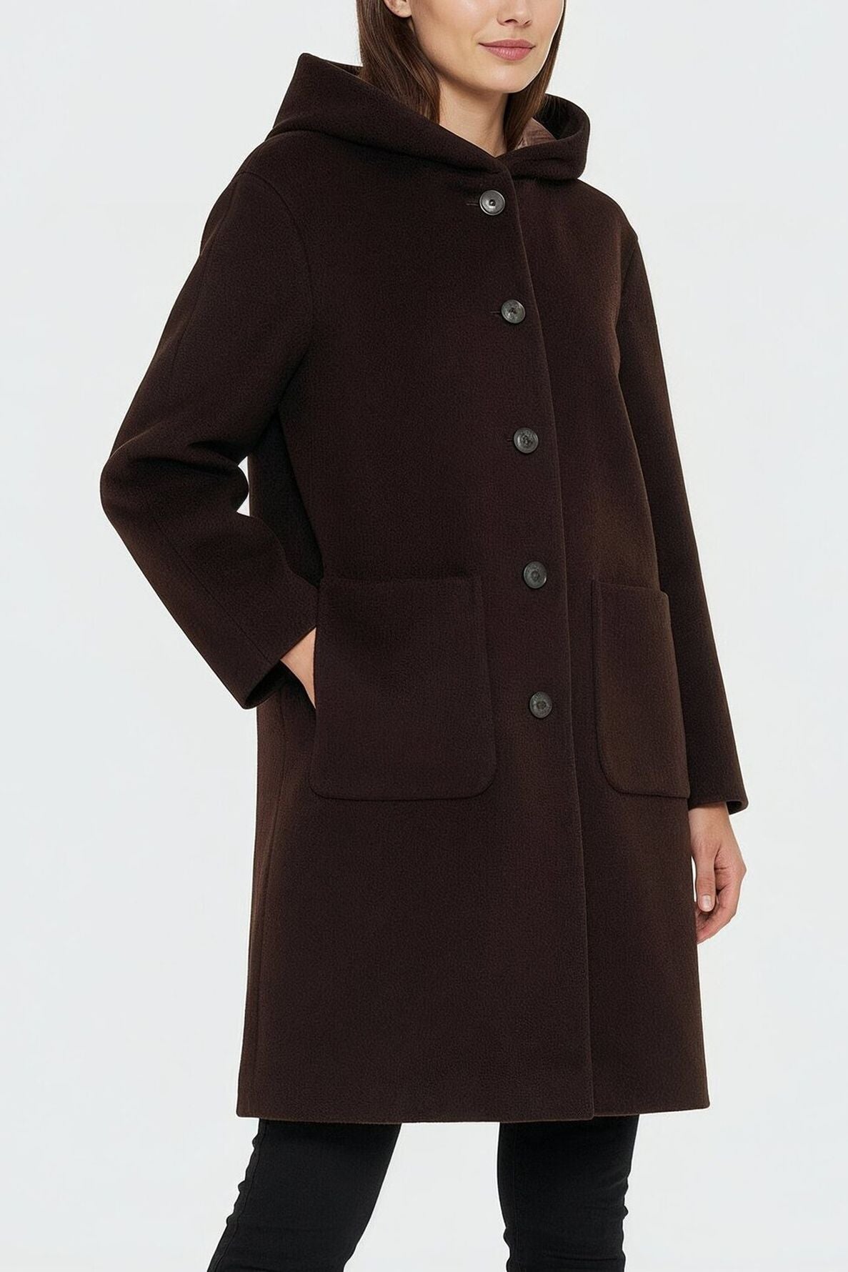 A woman wearing a dark brown manteau à capuche with large buttons and two front pockets stands with one hand in her pocket against a plain light background.