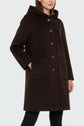 A woman wearing a dark brown manteau à capuche with large buttons and two front pockets stands with one hand in her pocket against a plain light background.