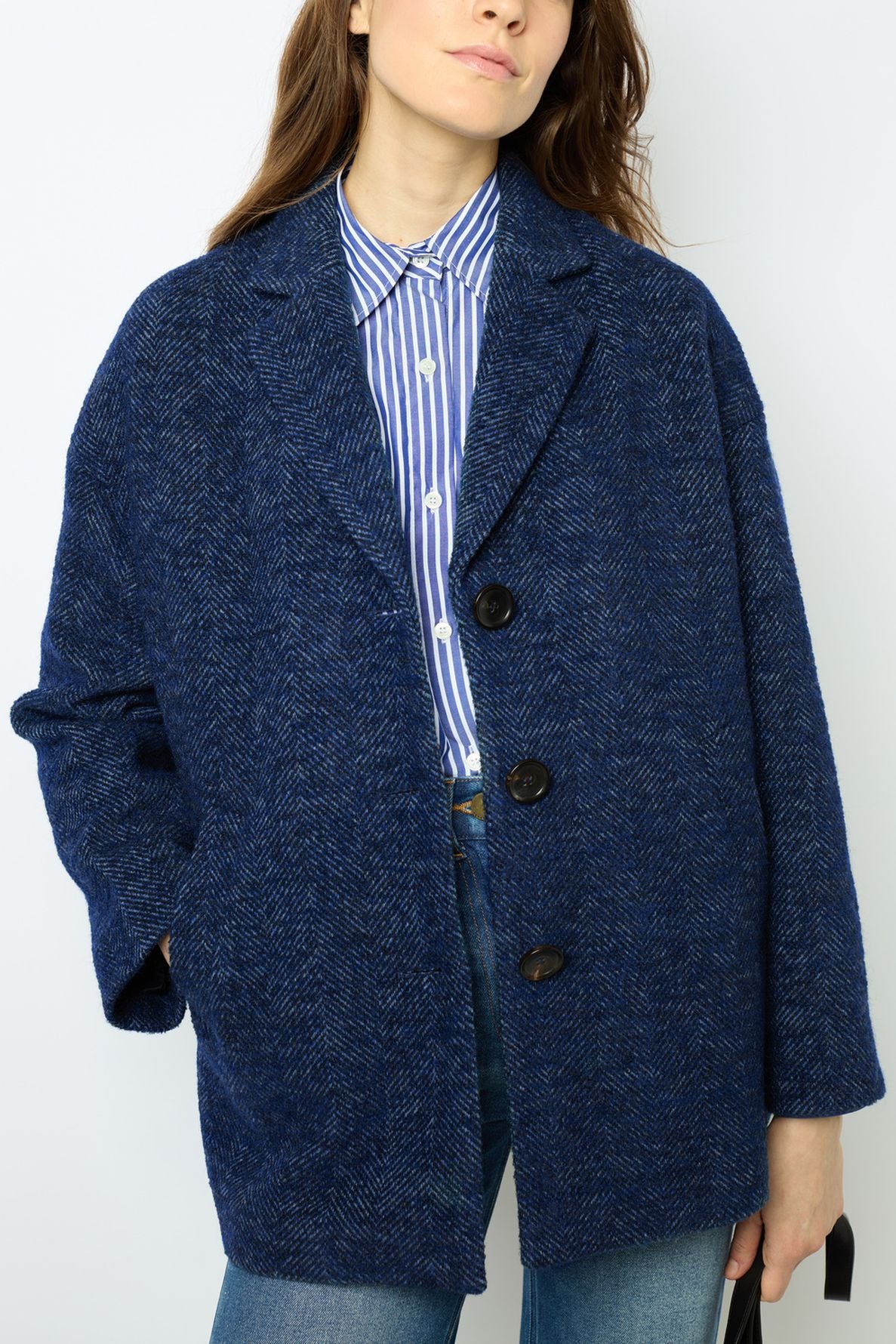 A person wearing a blue manteau laine textured coat over a blue-and-white striped shirt with jeans, standing with one hand in their pocket against a plain background.