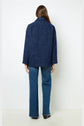 A person with long brown hair, seen from behind, wears a manteau court oversize in dark blue and blue jeans with black shoes, standing against a plain white background.