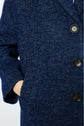 A person wearing a blue manteau à chevrons herringbone-patterned coat with large black buttons stands with one hand in the pocket.