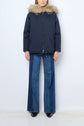 Person wearing a navy parka courte coton with a capuche fourrure amovible, blue wide-leg jeans with a darker denim stripe, and black shoes, standing against a plain white background.