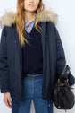 A woman wearing a navy parka courte coton with a capuche fourrure amovible, a blue v-neck sweater over a striped shirt, and blue jeans, holding a black leather handbag.