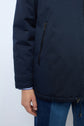 A person wearing a dark blue parka courte coton with a zipped pocket, a blue striped shirt cuff showing, and blue jeans. Only the lower half of the jacket and one hand are visible.