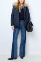 A person in a navy parka courte coton with a capuche fourrure amovible, blue sweater over a striped shirt, flared blue jeans, black ankle boots, and holding a black handbag stands against a plain background.