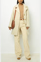 A woman wearing a manteau mi-long cream coat, a beige sweater, wide-leg cream pants, and tan platform shoes, with a brown shoulder bag, stands against a white background.
