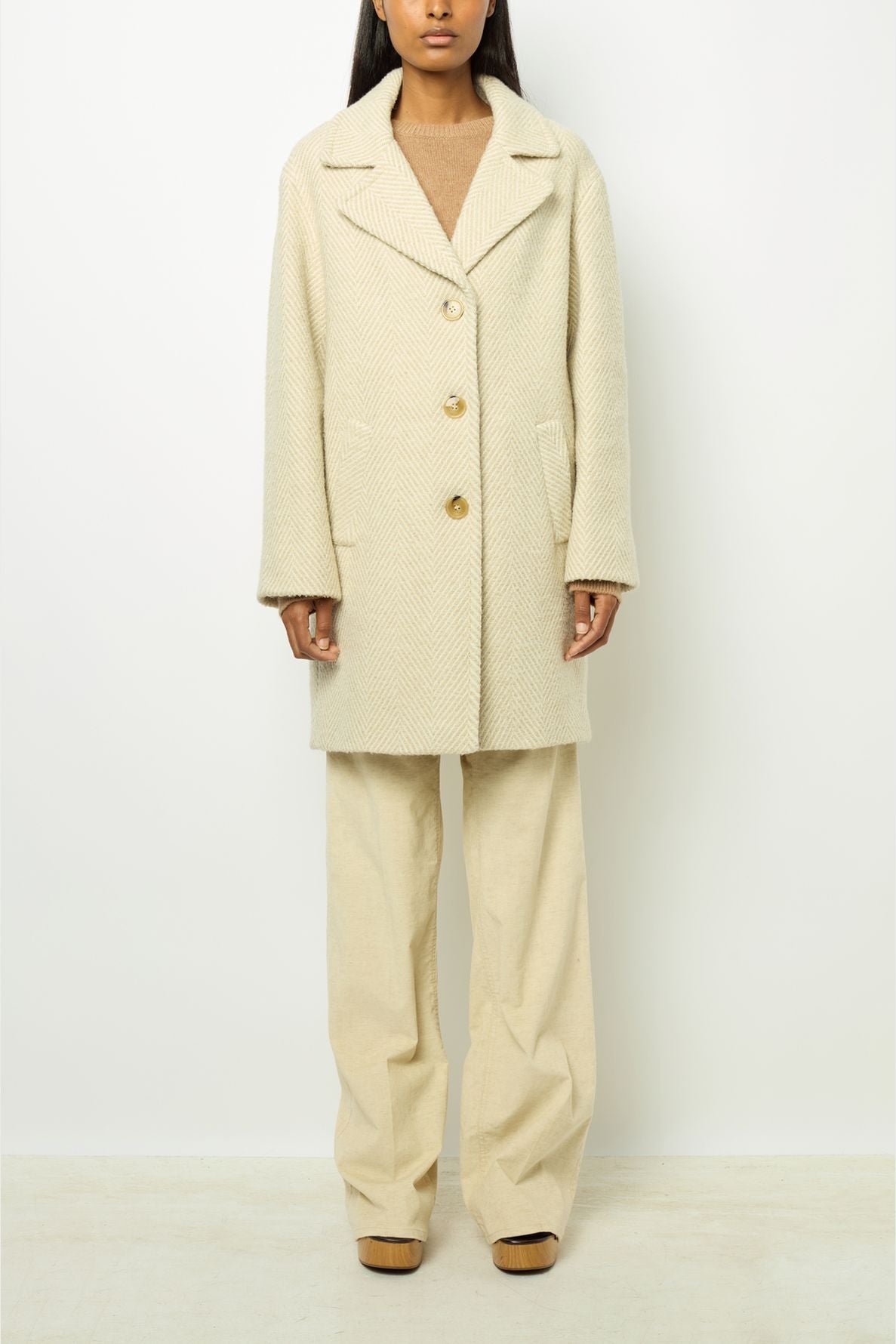 Person wearing a cream-colored manteau mi-long with large buttons over a beige sweater and wide-leg pants, standing against a plain white background.