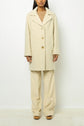 Person wearing a cream-colored manteau mi-long with large buttons over a beige sweater and wide-leg pants, standing against a plain white background.