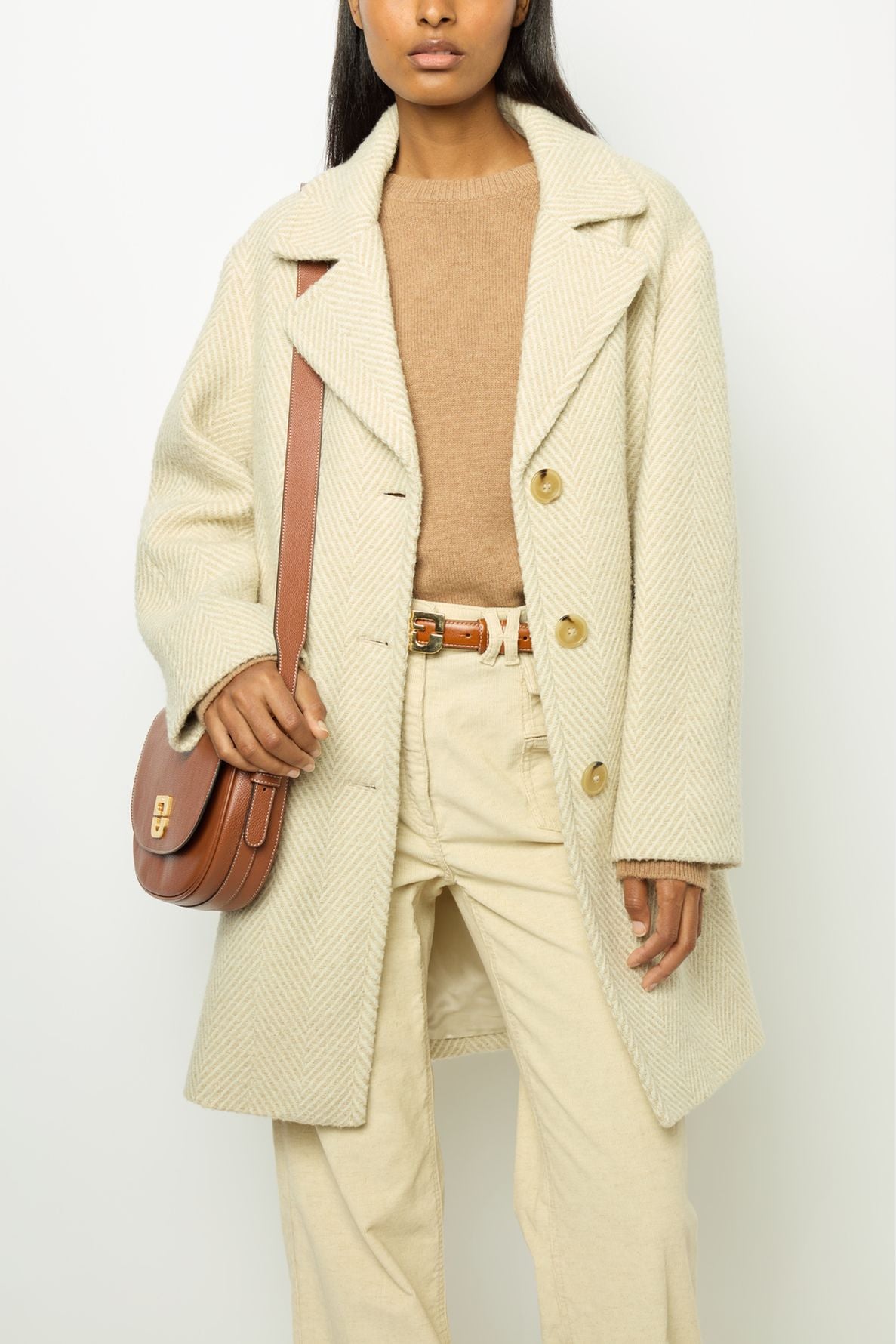 Woman wearing a cream manteau laine textured coat, beige sweater, off-white pants, and carrying a brown crossbody bag. Her hand rests on the bag’s strap, and she is photographed from mid-face down.