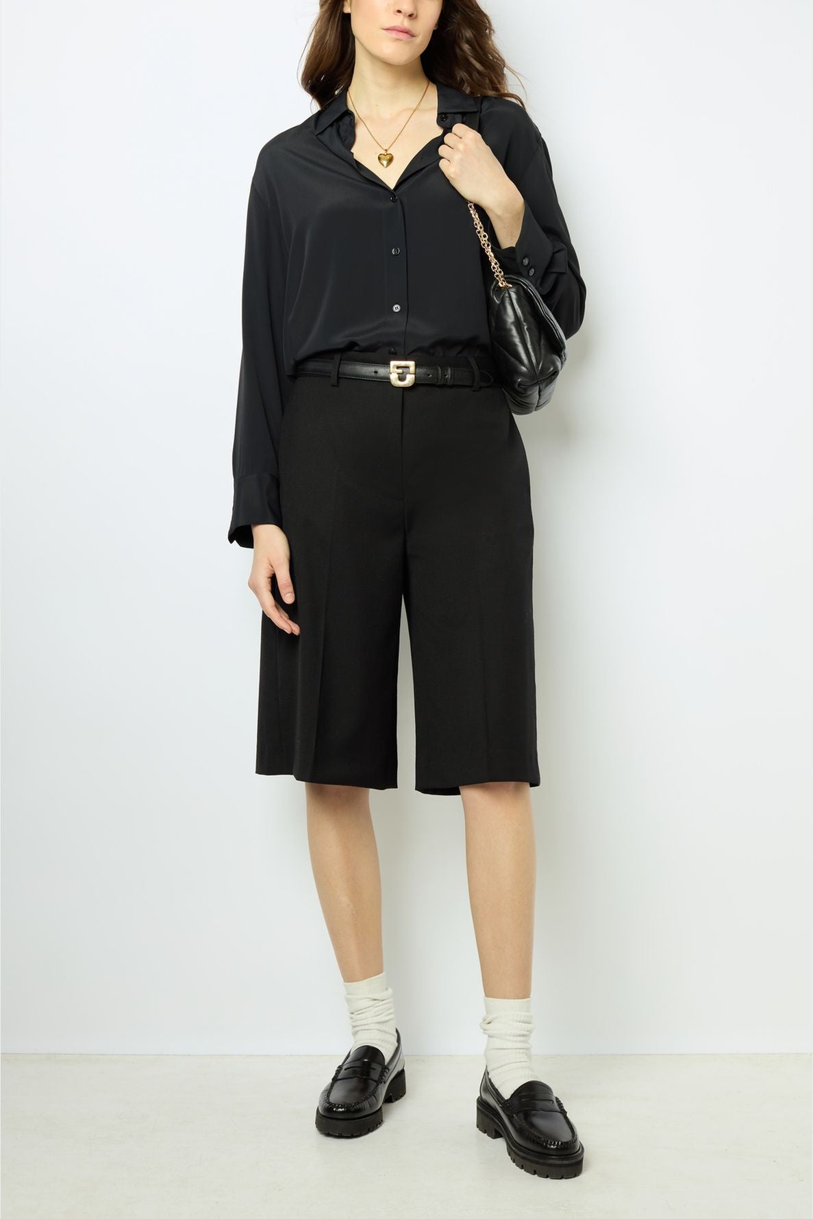 A person wearing a black button-up shirt, knee-length black shorts, black loafers with white socks, a black belt, layered necklaces, and a black shoulder bag stands against a plain white background.