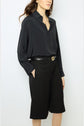 A woman wearing a black button-up blouse tucked into knee-length black shorts with a belt, standing against a plain light background. Her hands are in her pockets and her hair is loose.