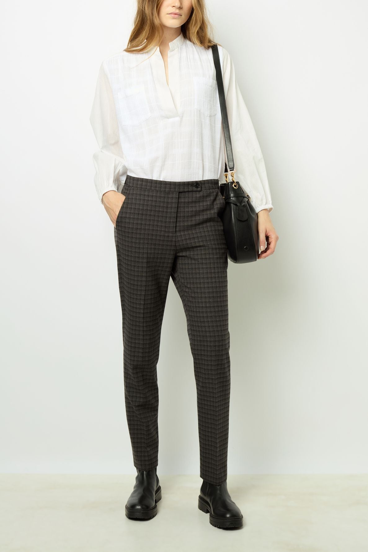 A person stands against a white wall wearing a white long-sleeve blouse, dark pantalon à carreaux coupe droite, black boots, and holding a black handbag.