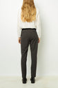 A person with long wavy hair, wearing a white blouse and dark pantalon à carreaux coupe droite, stands facing away from the camera against a plain white background.