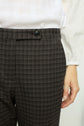 A person wearing a white textured blouse and a pantalon à carreaux coupe droite with a black button stands against a light background. Only their torso and right hand are visible.