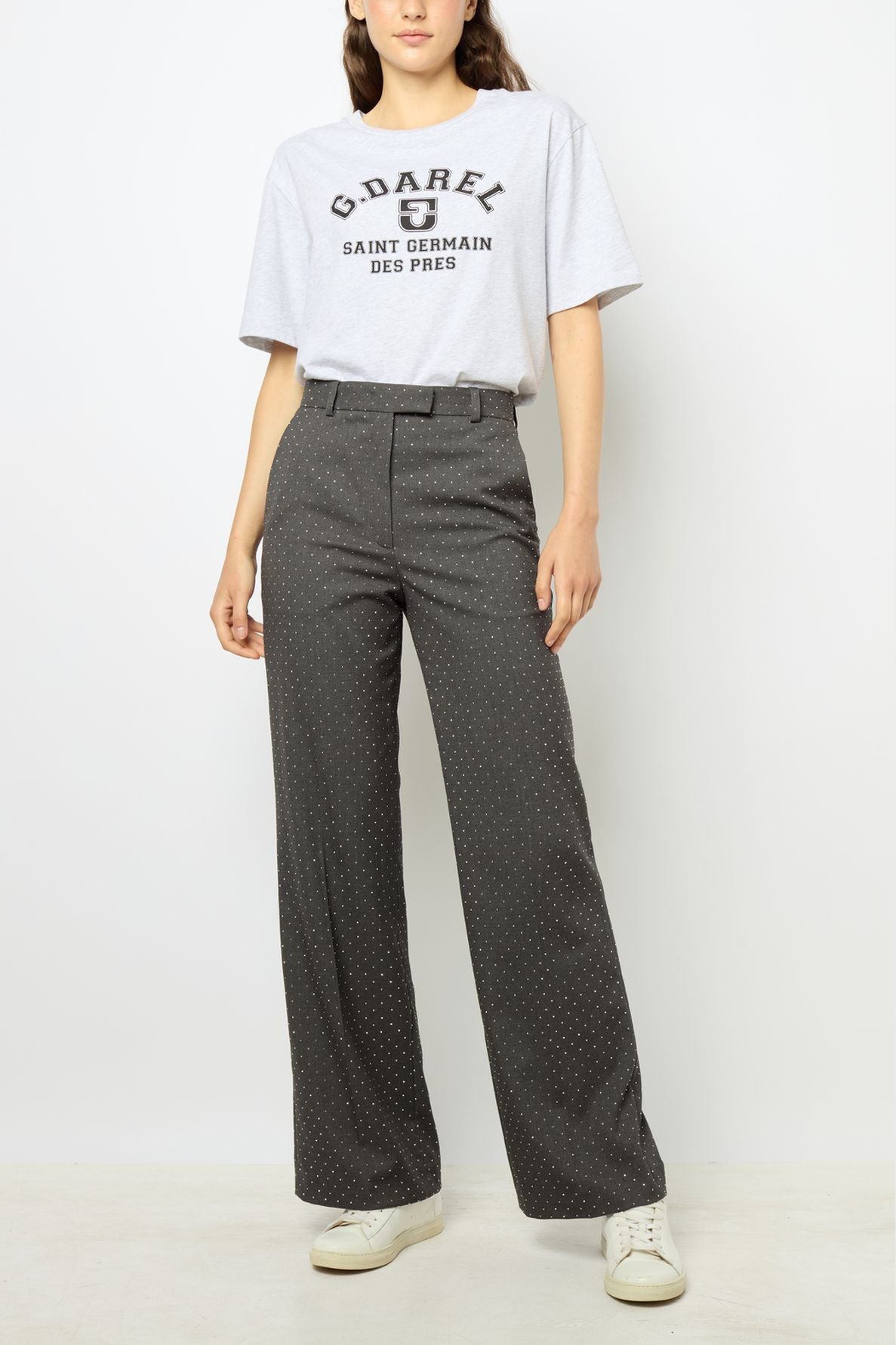 A woman stands against a plain background wearing a light gray graphic t-shirt, high-waisted dark gray polka dot pantalon droit from the collection Studio, and white sneakers. Her right hand is relaxed by her side.