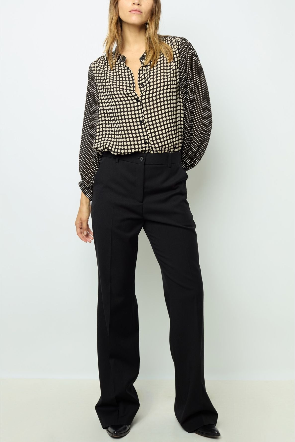 Woman wearing a black and white checkered blouse with long sleeves and black high-waisted wide-leg trousers, standing against a plain light background.