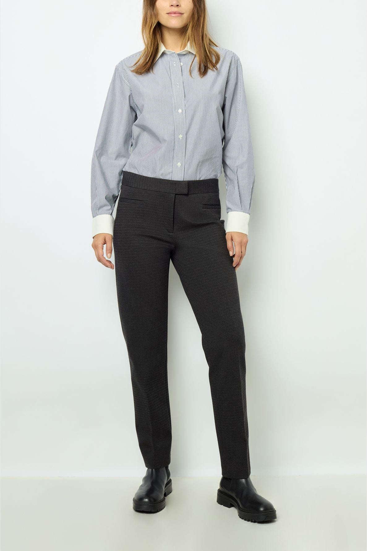 A person wearing a blue button-up shirt, black boots, and a pantalon droit 7/8 en matière stretch with un motif pied de puce stands against a plain white background. Their face is partially out of frame.
