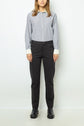 A person stands against a plain background wearing a blue-striped button-down shirt with white cuffs, pantalon droit 7/8 in dark tailored fabric, and black shoes. Their hair is down and their hands are relaxed at their sides.